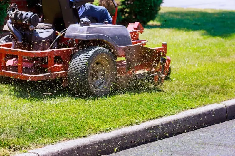 Commercial zero-turn mower cutting a green lawn along a street curb